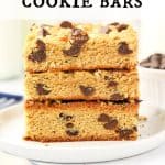 Stack of three thick peanut butter cookie bars with chocolate chips and chopped peanuts on a white plate. Dish of chocolate chips in the background. Text overlay reads: Peanut Butter Cookie Bar.