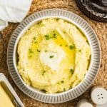 A bowl of Boursin Cheese Mashed Potatoes is served on a dinner plate with melted butter pooling in the center and garnished with fresh parsley on top. The table setting includes a butter dish with a knife, a white napkin, a stack of brown and white dishes with silverware, and salt and pepper shakers nearby.