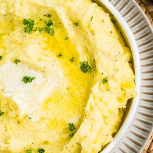 Creamy Boursin Mashed Potatoes served in a pretty gray bowl, garnished with melted butter and fresh parsley. The mashed potatoes are smooth and fluffy with visible flecks of black pepper and herbs, showcasing a rich and buttery texture.