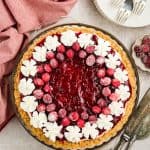 Whole Cranberry Cheesecake in a pie dish. The cheesecake is garnished with piped whipped cream 'stars' around the outer edge, and then sugared cranberries just inside the whipped cream circle. Plates and forks are in the background, serving utensils are off to the side.