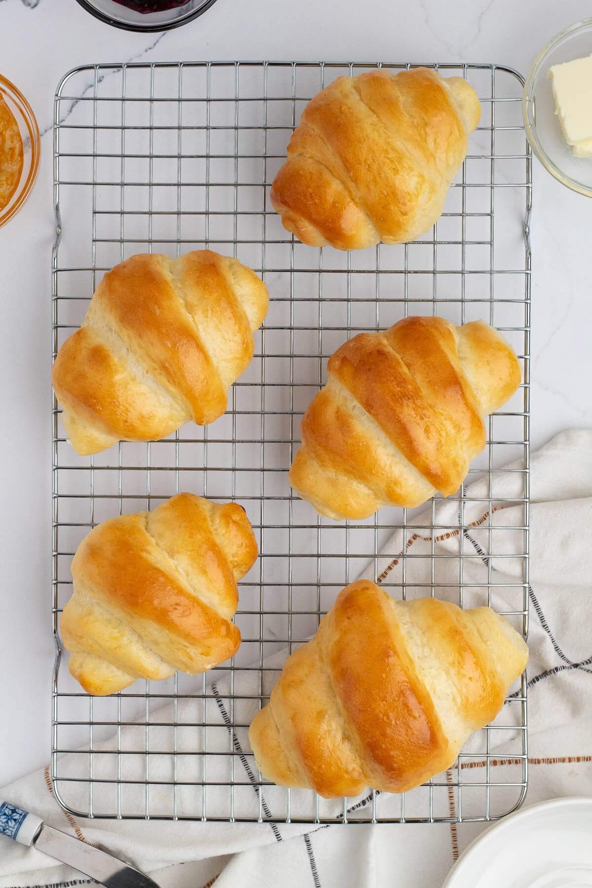 Five golden crescent rolls cooking on a baking rack. There is a linen underneath the front corner of the rack and some preserves and butter surrounding the baking rack.