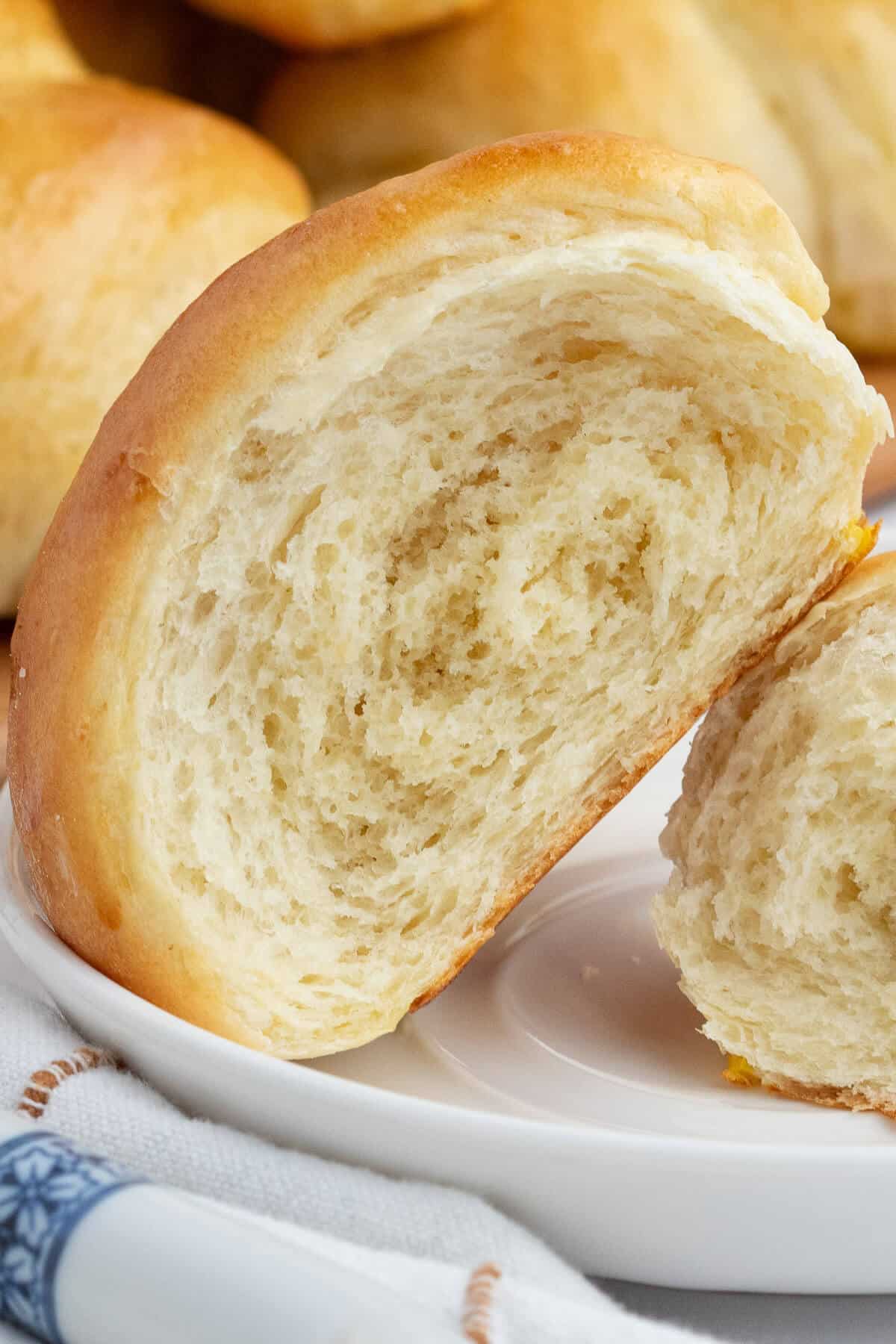 Crescent roll torn in half showing the soft texture inside, leaning on the other half in which only a corner is visible. Roll is on a white plate and there are more rolls in the background.