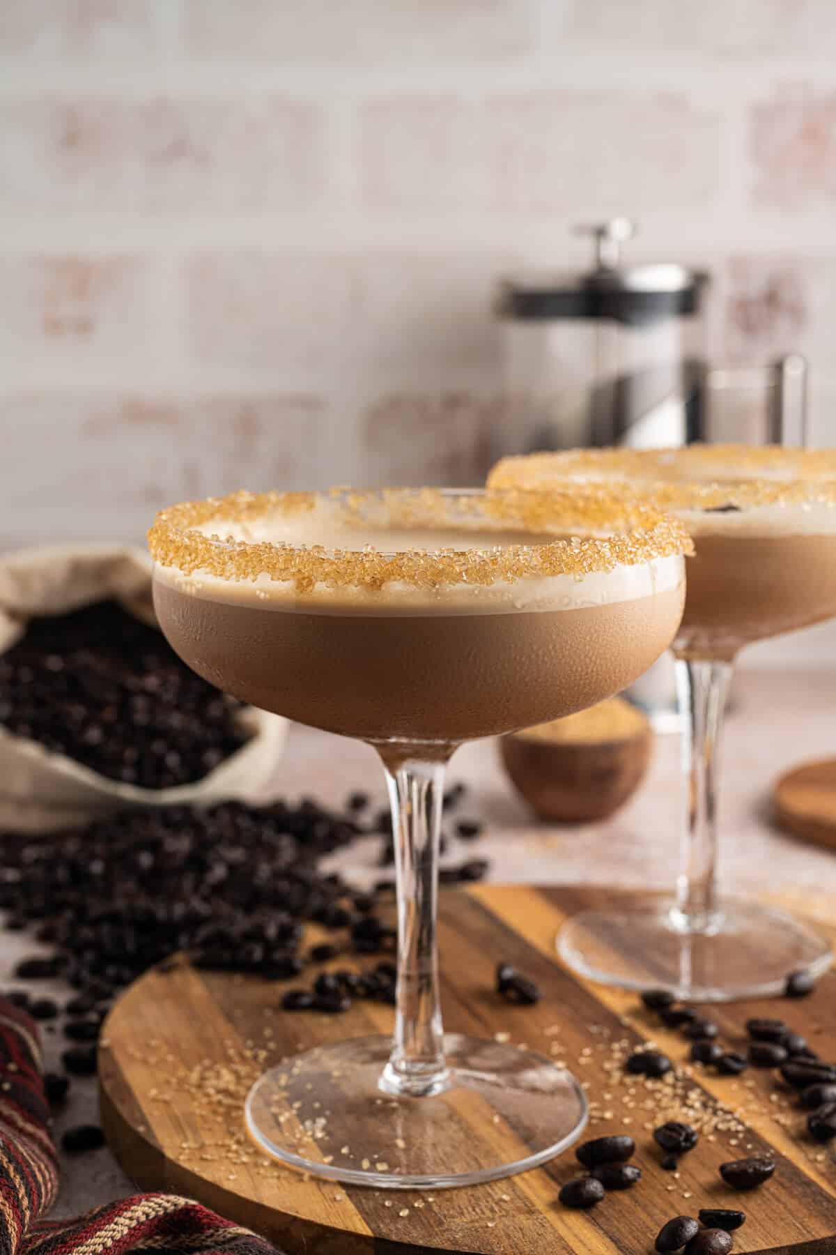 Espresso martini mocktail in a coupe glass with a golden raw sugar rim and frothy top, styled with scattered coffee beans and a bag of whole beans in the background.