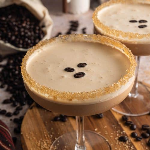 Espresso martini mocktail served in a coupe glass with a raw sugar rim, topped with three coffee beans on a creamy frothy layer, surrounded by scattered coffee beans on a wooden board.