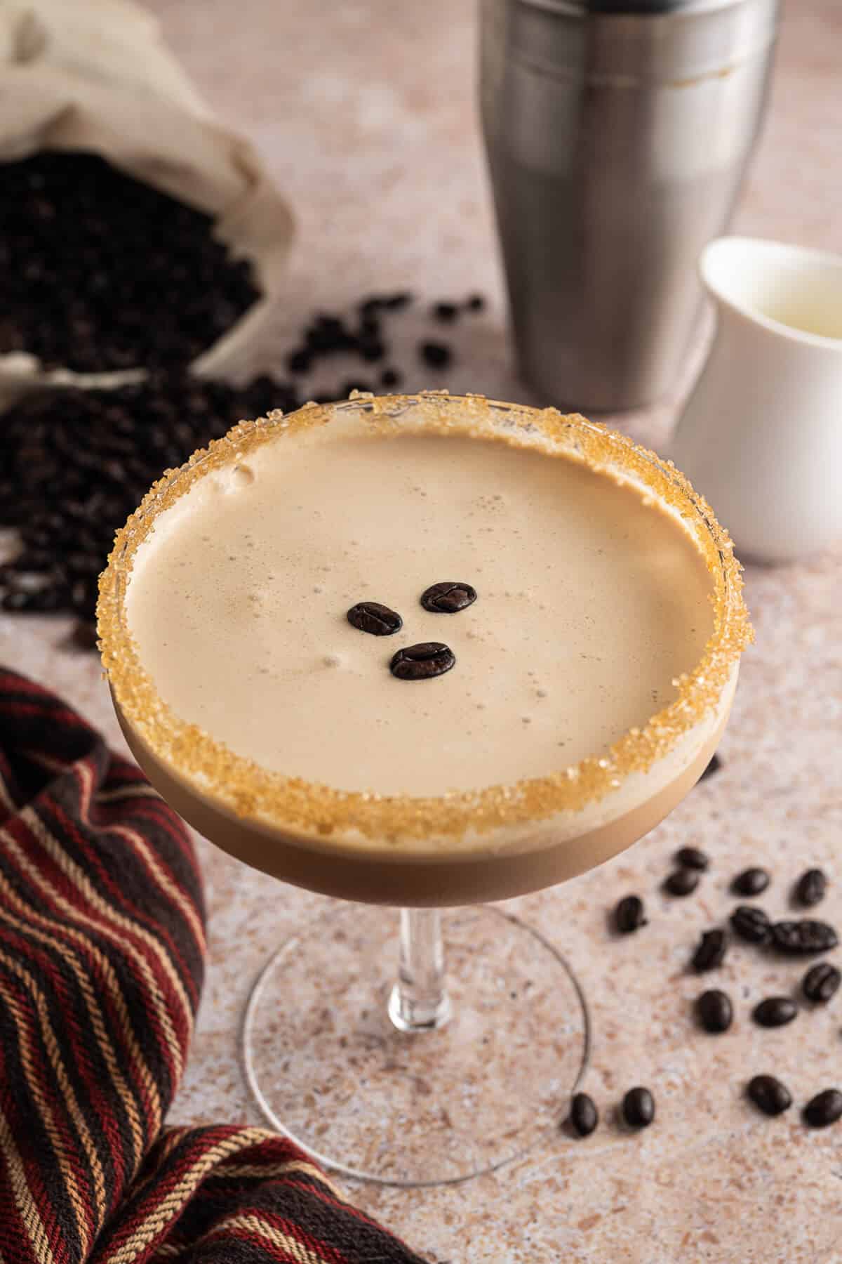 Espresso martini mocktail in a coupe glass with a sugared rim and frothy top, garnished with three coffee beans. A cocktail shaker, cream pitcher, and scattered coffee beans are in the background.