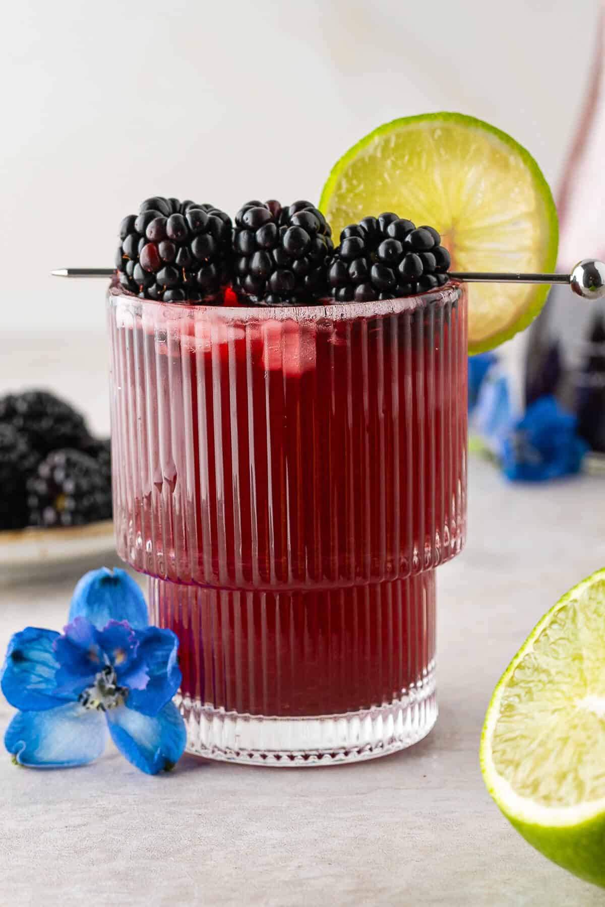 Blackberry Mocktail in a highball glass garnished with fresh blackberries on a cocktail pick and a slice of lime. blackberries and lime in the background, blue flower at the bottom of the glass.