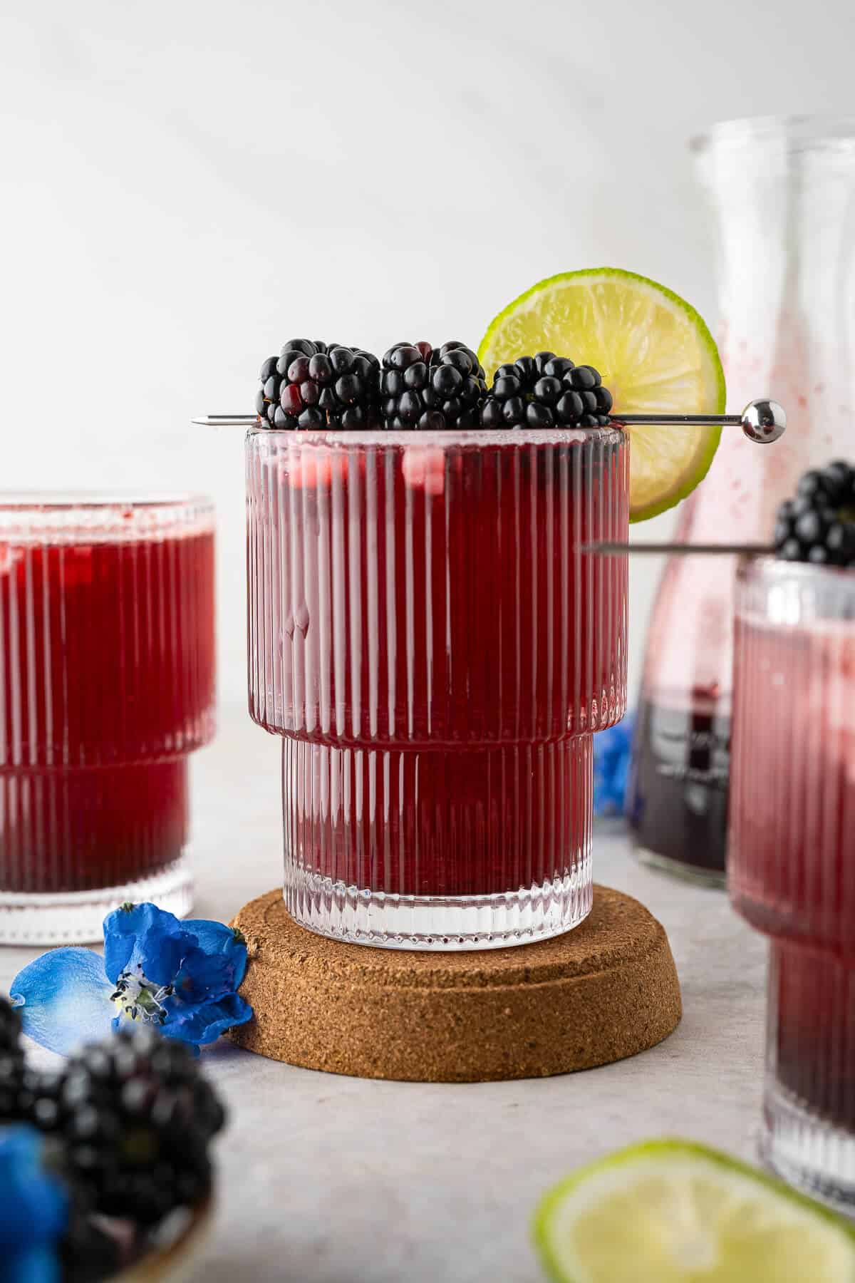 Blackberry Mocktail in a ribbed glass on a cork coaster, garnished with fresh blackberries and a lime slice, with more glasses and a carafe in the background.