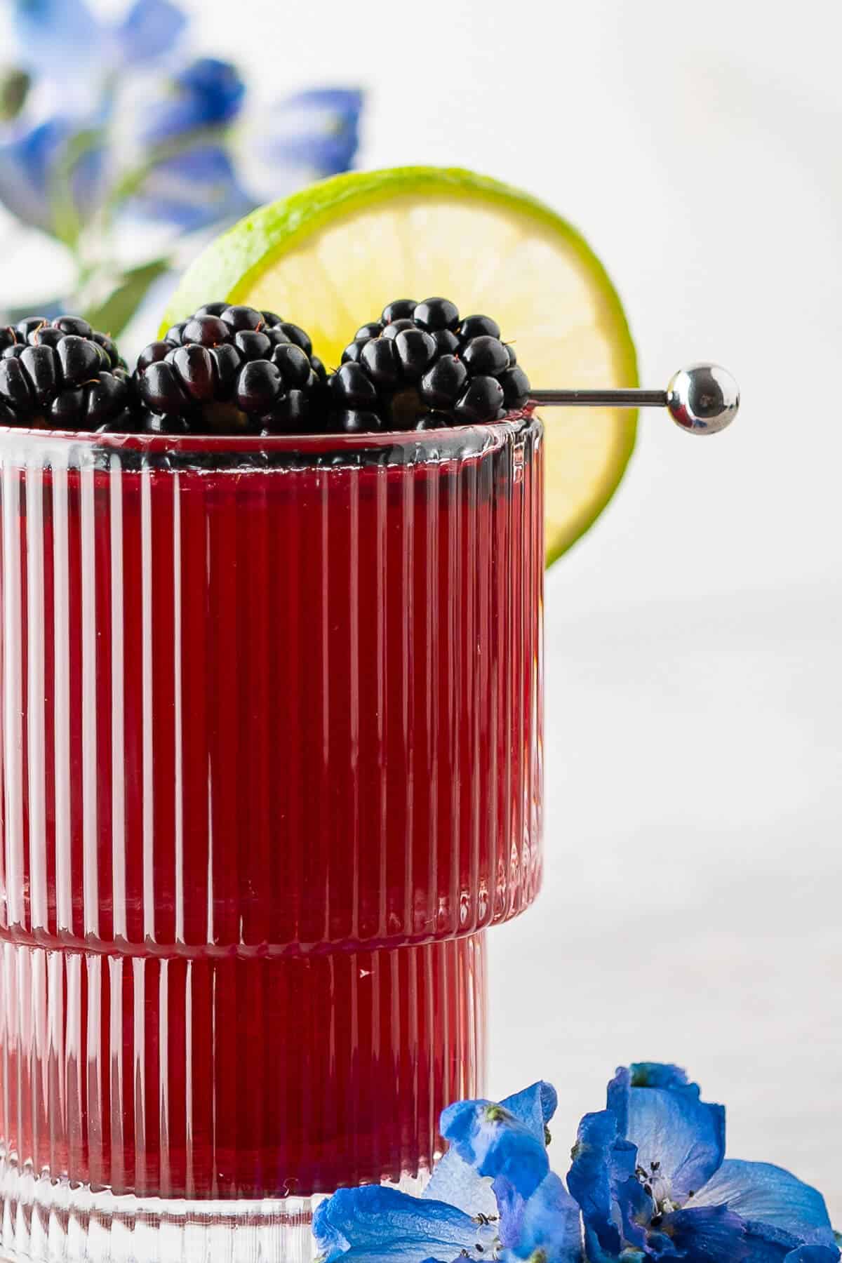 Close-up image of a blackberry mocktail in a ribbed glass, garnished with blackberries on a cocktail pick and a lime slice, with blue flowers beside the glass.