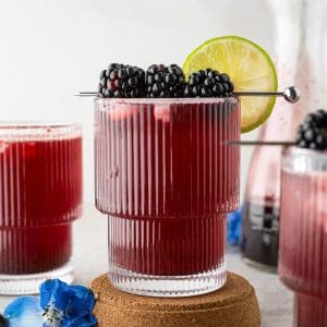 Blackberry Mocktail in a ribbed glass on a cork coaster, garnished with fresh blackberries and a lime slice, with more glasses and a carafe in the background.