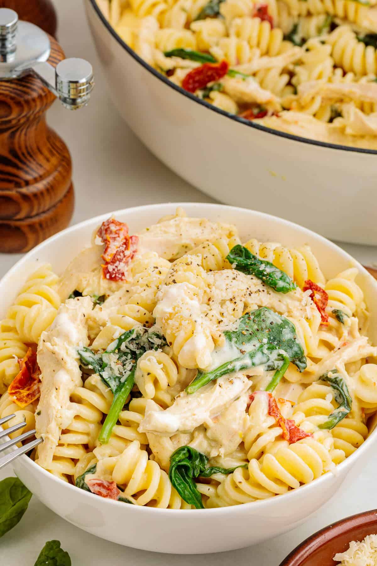 White bowl of Boursin Chicken Pasta with sun-dried tomatoes and spinach. Pot of pasta in the background.