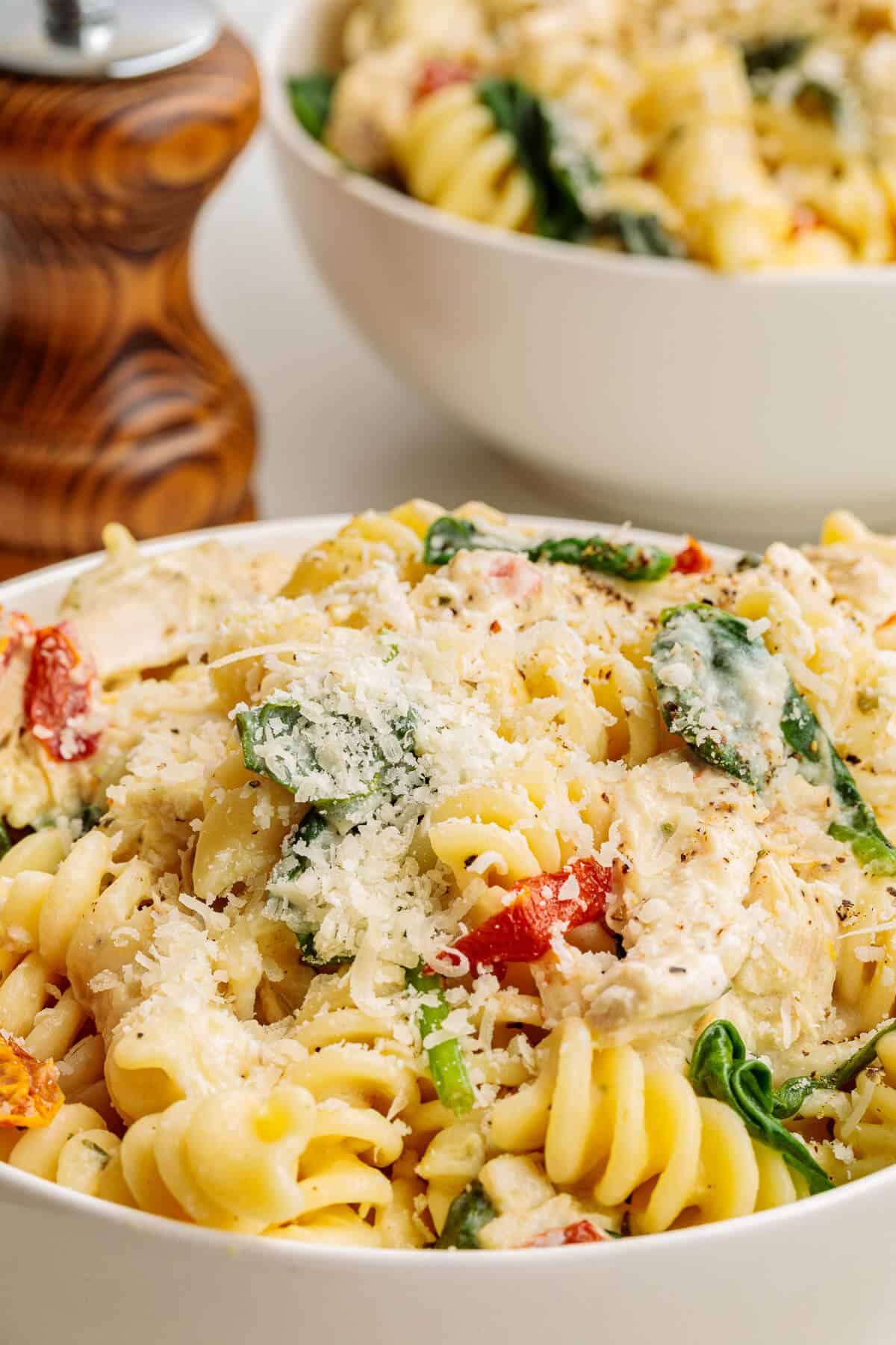 Two bowls of Boursin Chicken Pasta, one in the background. Pasta is in a creamy sauce and has chicken, sun-dried tomatoes and spinach, garnished with parmesan cheese and fresh ground pepper.