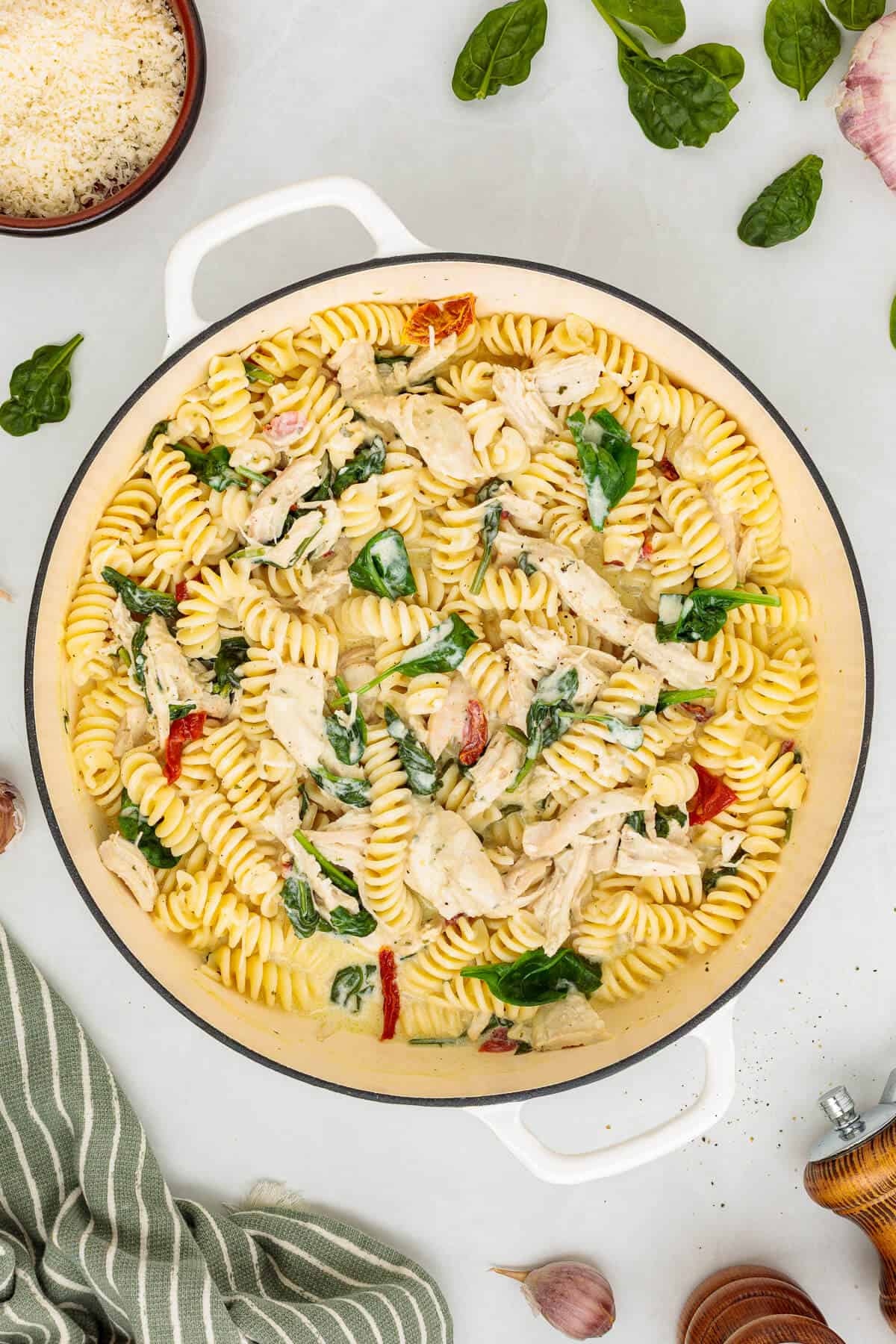 Overhead view of a white dutch oven filled with Pasta, chicken, sun-dried tomates and spinach in a creamy Boursin Cheese sauce. Green striped towel lays along the bottom left corner, grated parmesan cheese and spinach leaves fill the top corners.