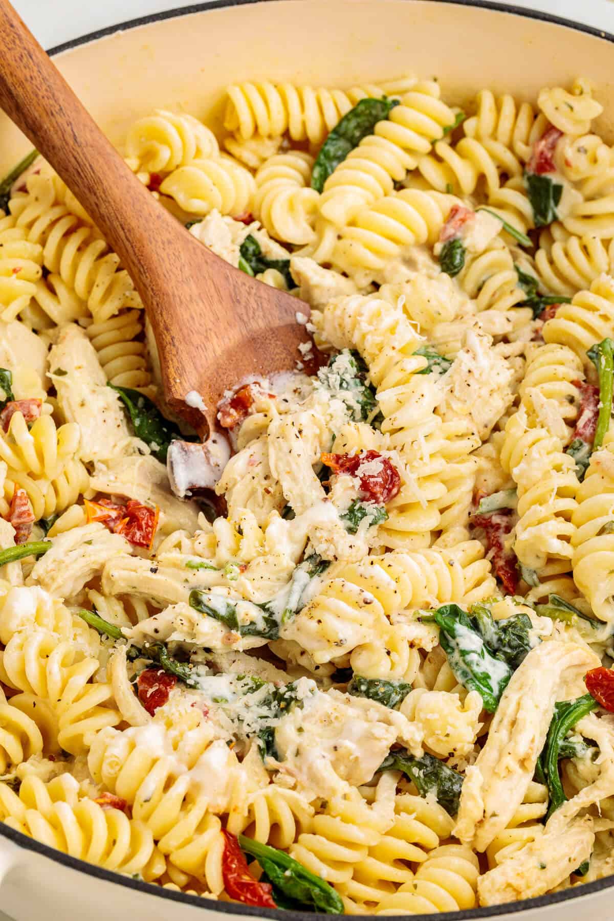 Large pot of Creamy rotini pasta in a boursin cheese sauce with spinach and sun-dried tomatoes, a wooden spoon is scooping into the pasta.