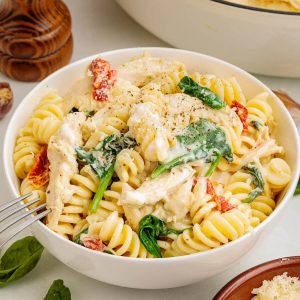 Bowl of rotini pasta with chicken, spinach and sun-dried tomatoes in a creamy Boursin cheese sauce, garnished with parmesan cheese and fresh ground pepper. Pot of pasta and pepper mill in the background, small bowl of grated parmesan cheese peeking out from the corner.