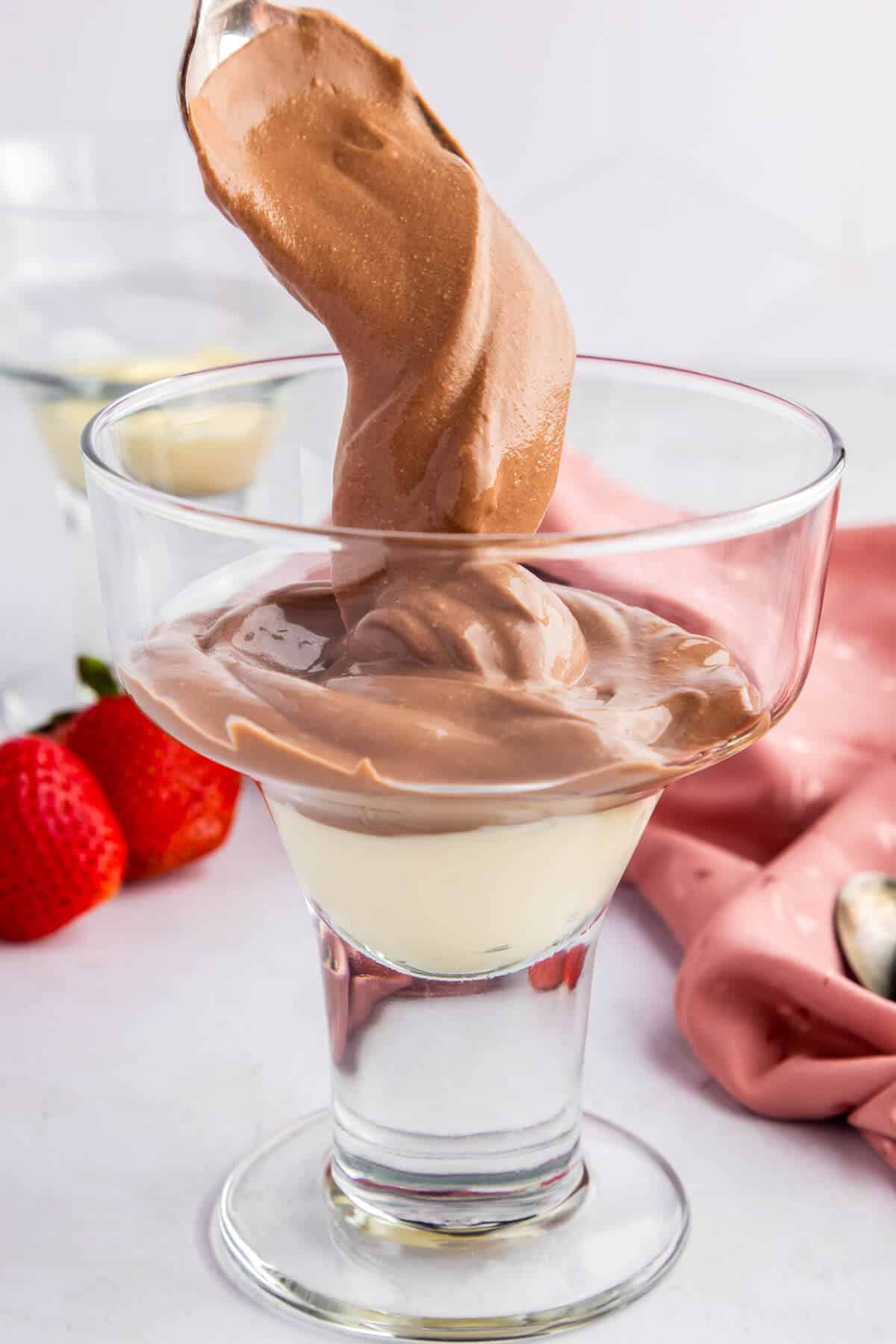 A spoon dropping a dollop of Cottage Cheese Chocolate Mousse into a dessert glass. Fresh strawberries in the back ground and a pink table linen.