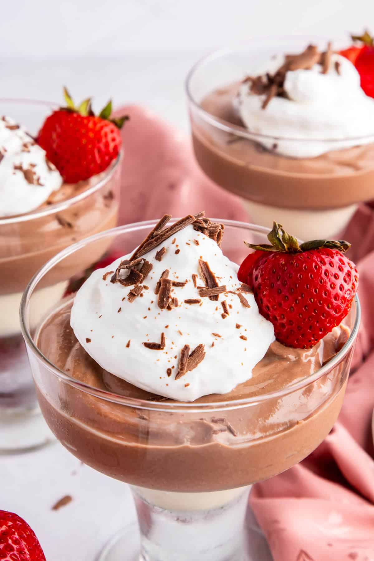 Three stemmed dessert dishes filled with Cottage Cheese Chocolate mousse, one in the front and two in the background. They are topped with whipped cream and chocolate shavings, and garnished with a sliced strawberry with the stem in tact.