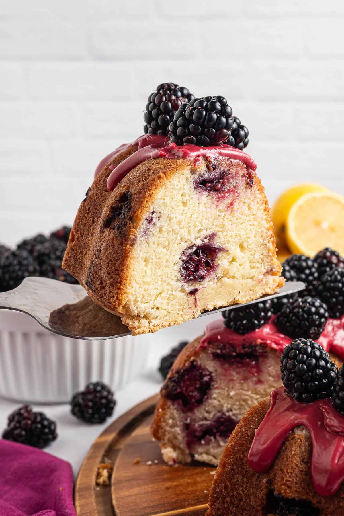 Slice of Lemon Blackberry Cake on a spatula, being held slightly above the whole bundt cake. The cake is topped with fresh juicy blackberries and a pink blackberry glaze.
