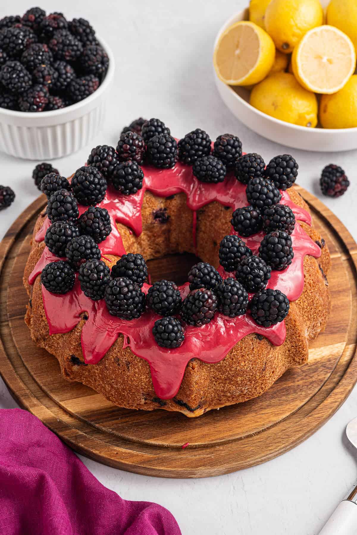 Whole Lemon Blackberry Bundt Cake on a round wooden board, topped with blackberry glaze and fresh juicy blackberries. A ramekin of fresh berries and a bowl of sliced lemons are in the background.