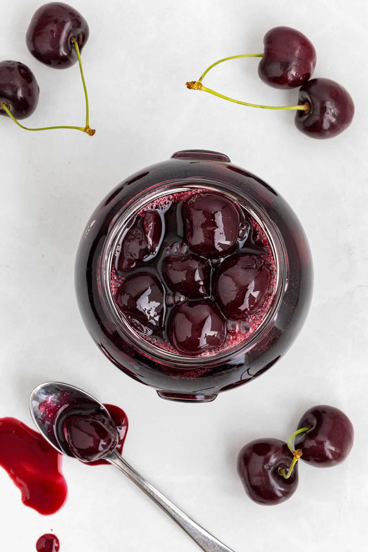 Jar of homemade cherry compote with whole cherries and syrup surrounded by fresh cherries. A spoon with compote on it is laying in the bottom left corner.