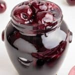 Glass jar full of Cherry compote in a thick glossy syrup. Whole cherries can be seen mounded on the top. A few fresh cherries surround the jar.