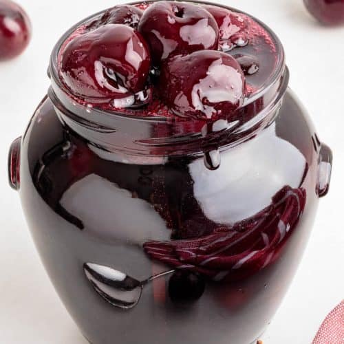 Glass jar full of Cherry compote in a thick glossy syrup. Whole cherries can be seen mounded on the top. A few fresh cherries surround the jar.