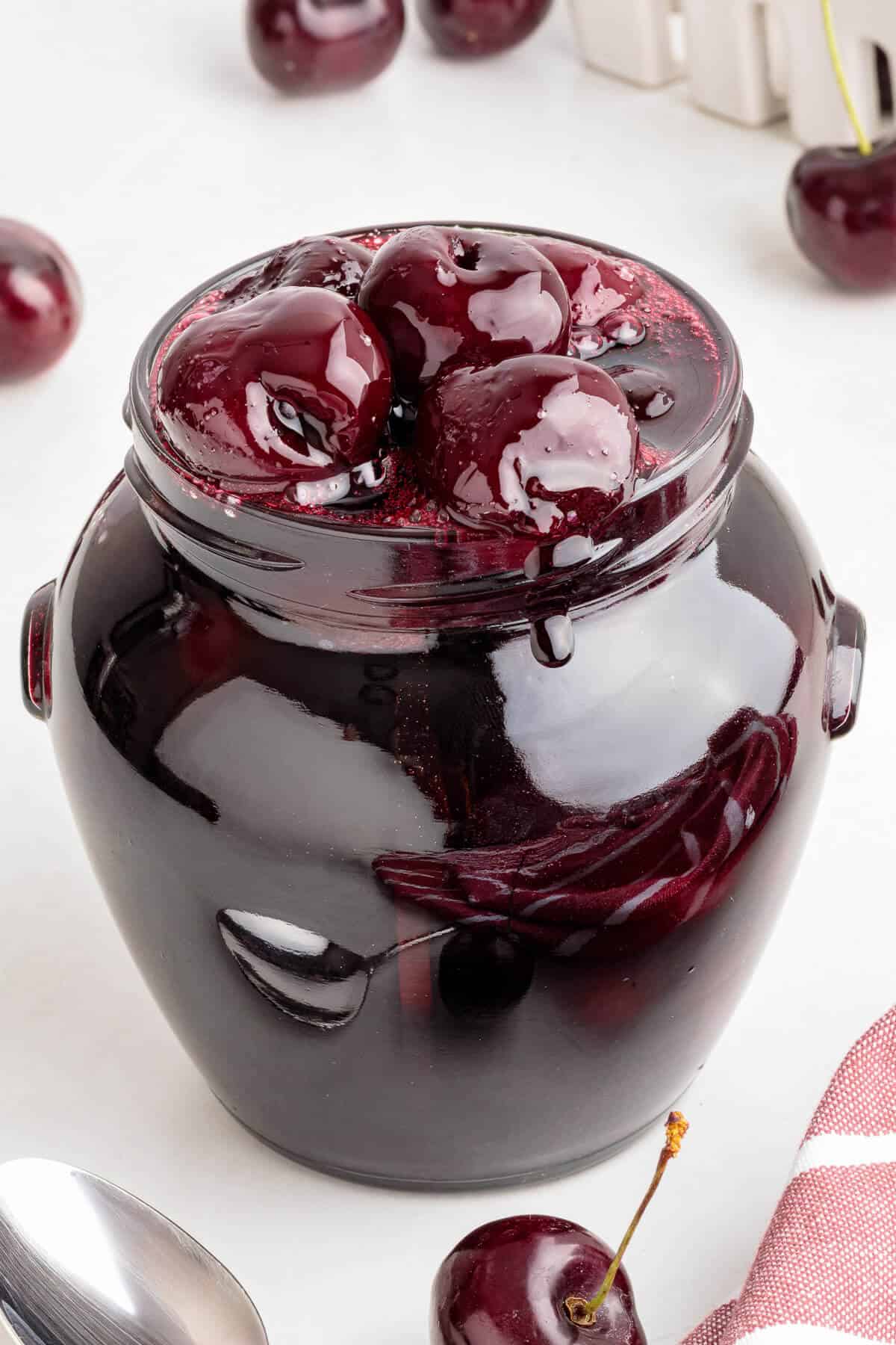 Glass jar full of Cherry compote in a thick glossy syrup. Whole cherries can be seen mounded on the top. A few fresh cherries surround the jar.