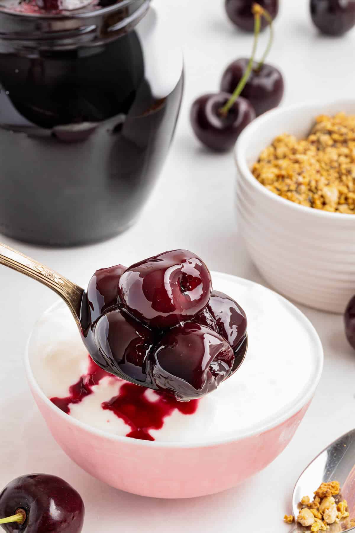 Spoon full of homemade cherry compote being placed onto a bowl full of yogurt. Granola and a jar of compote sit in the background.