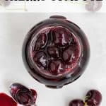 Glass jar full of homemade cherry compote. Fresh cherries surround the jar and a spoon with cherry compote on it is in the bottom left corner of the image. Text overlay reads: Cherry Compote