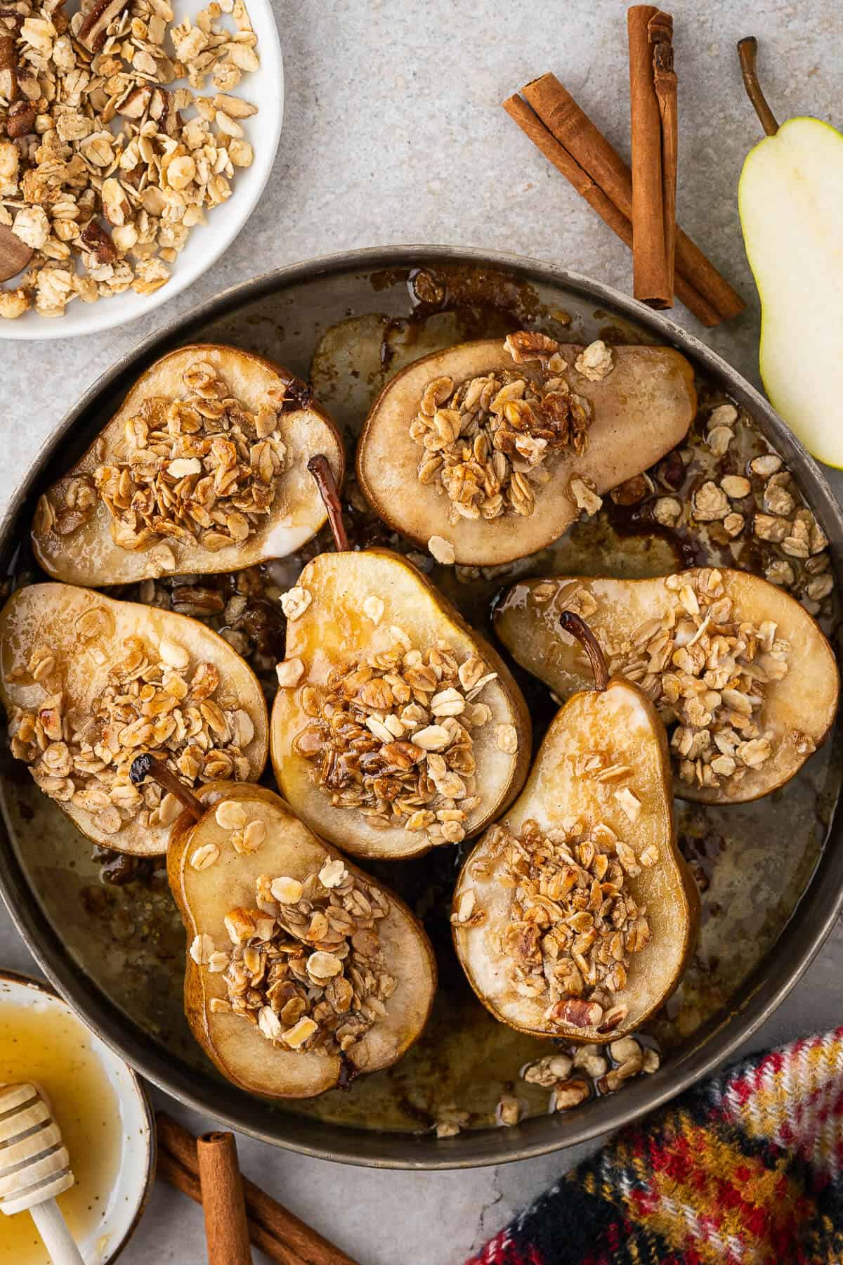 Baked pear halves topped with an oat crumble in a round baking dish, with cinnamon sticks, honey, granola, and a fresh pear around the dish.