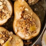Baked Pears in a round baking dish with just the bottom corner of the pan showing. The pears are topped with a sprinkle of granola and garnished with a piece of candied ginger.