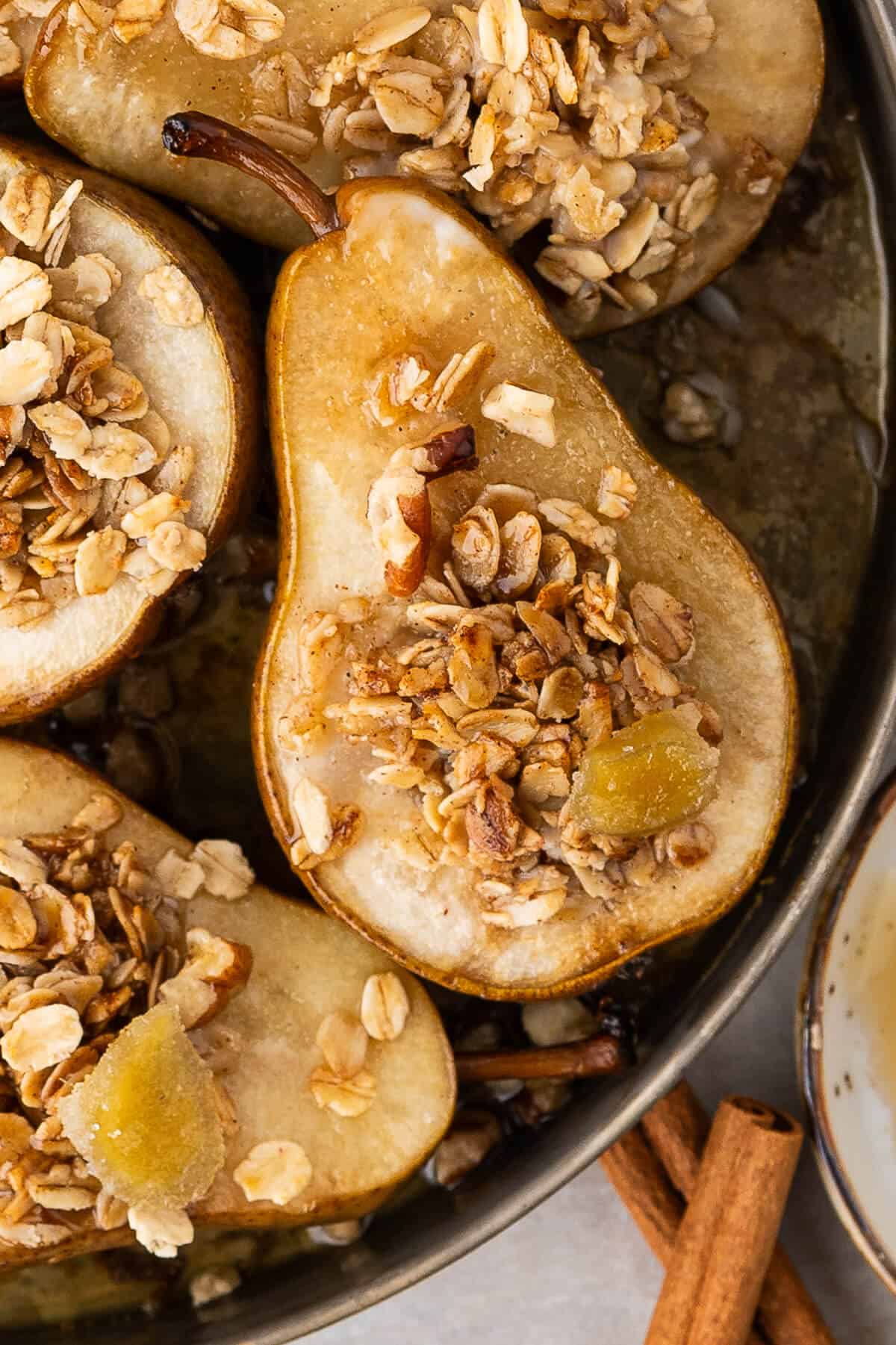 Baked Pears in a round baking dish with just the bottom corner of the pan showing. The pears are topped with a sprinkle of granola and garnished with a piece of candied ginger.