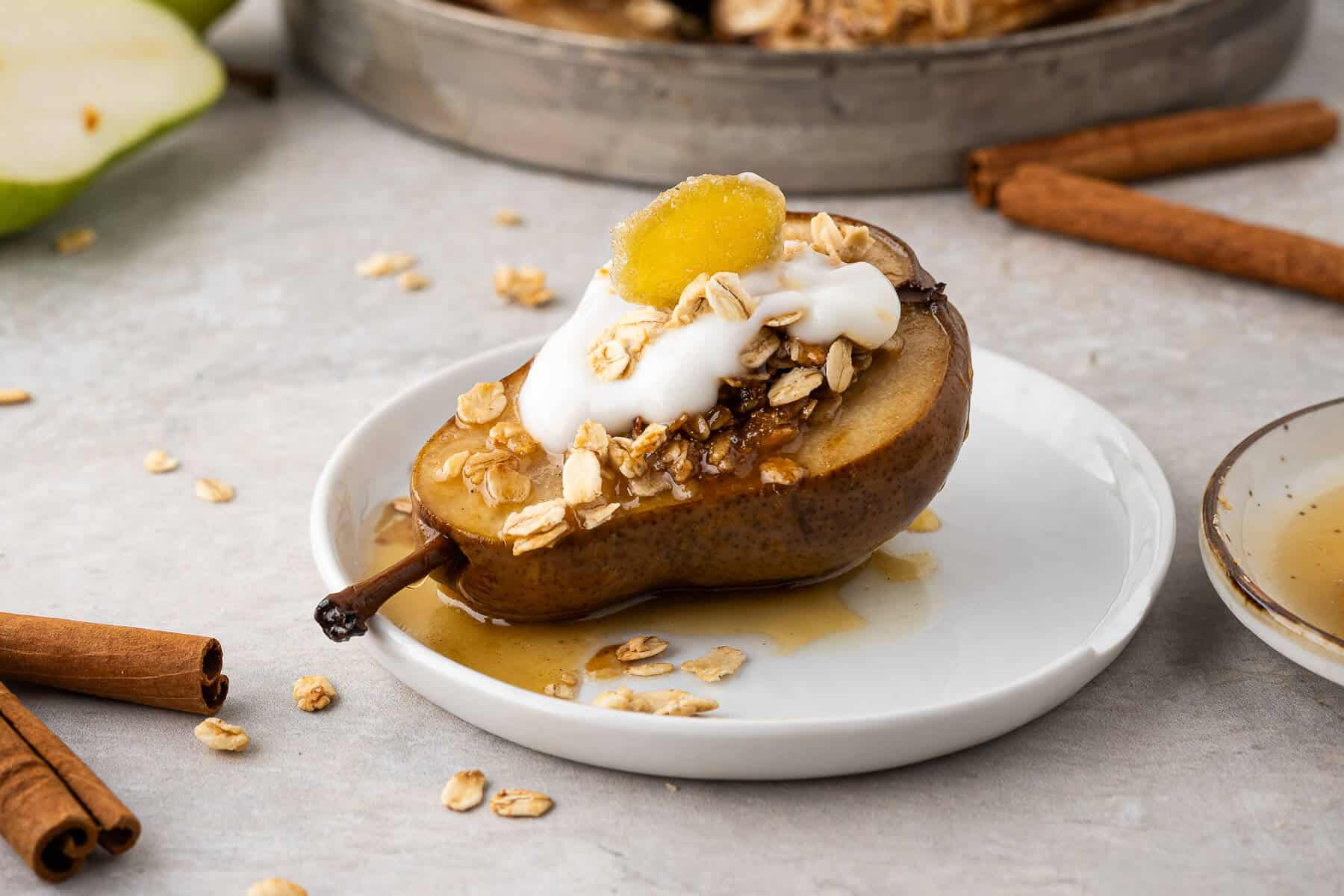 A baked pear half on a small white plate. It's topped with a honey ginger oat crumble, a dollop of vanilla yogurt, and garnished with a piece of candied ginger. Baking pan is in the back ground, oats and cinnamon sticks are scattered around.